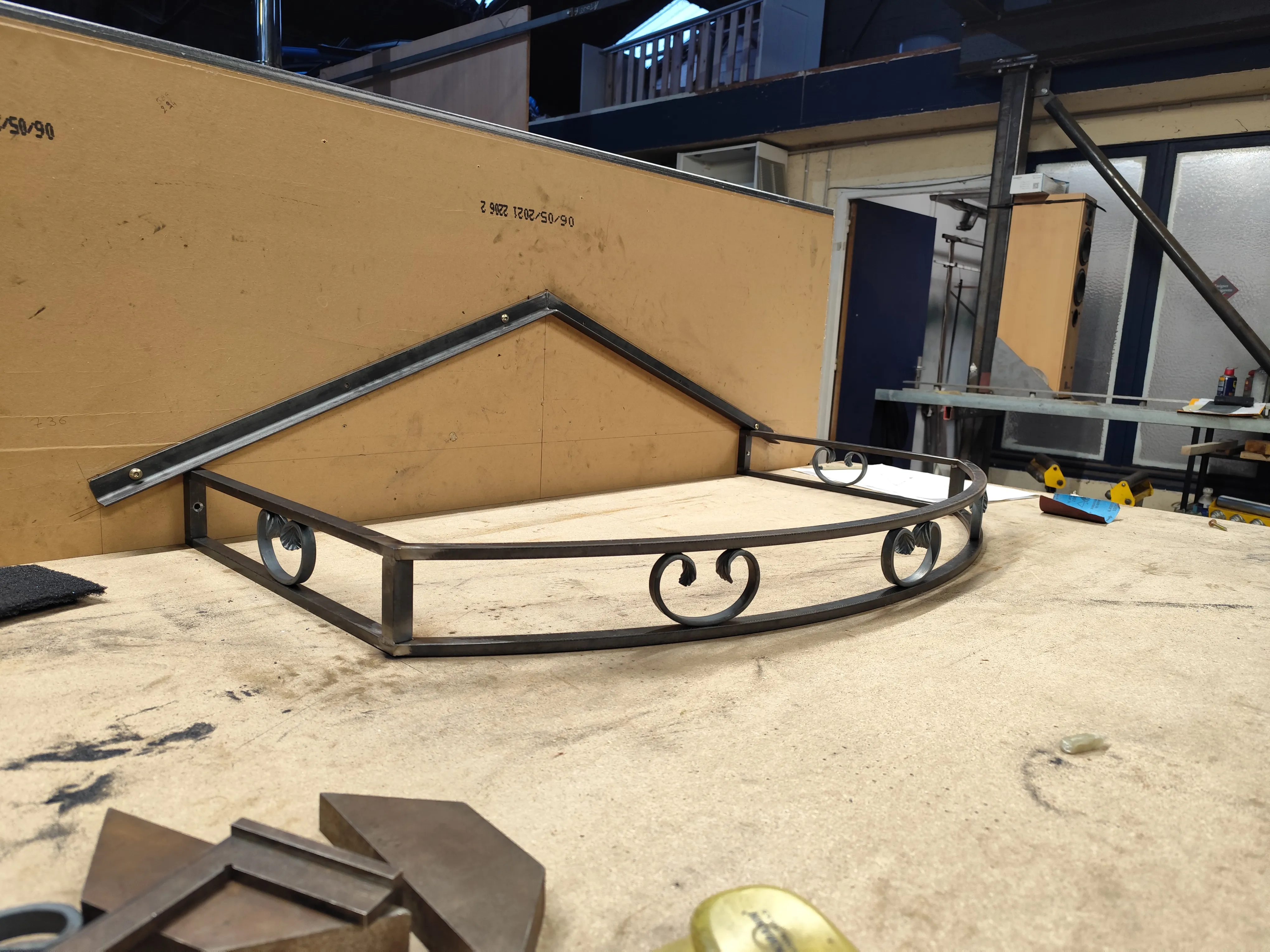 Vue en perspective de l'armature d'une marquise en acier sur mesure, montrant la façade cintrée avec ses motifs décoratifs en fer forgé sur un plan de travail.