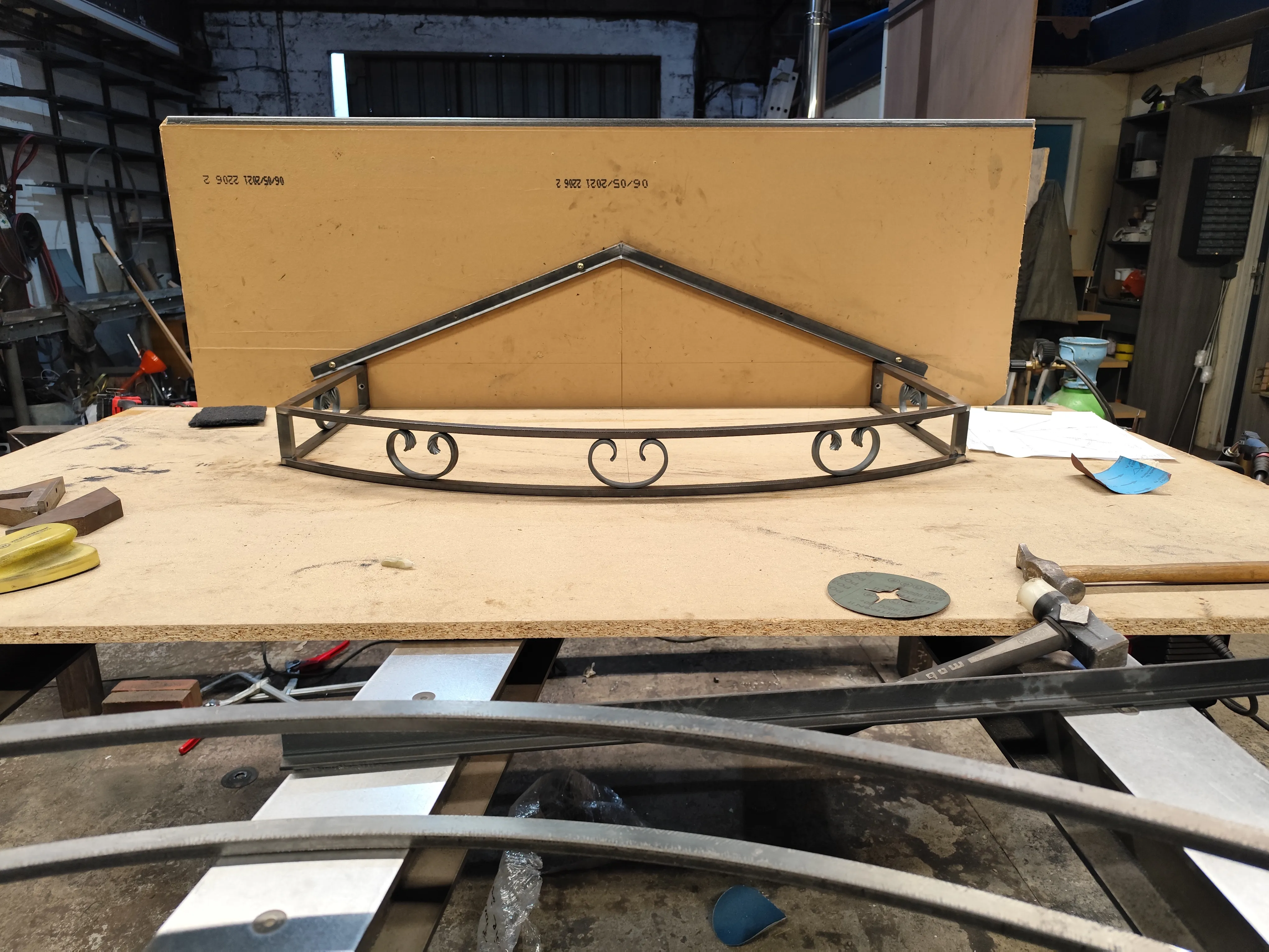 Armature de toit d'une marquise en acier de forme triangulaire avec une face avant courbée ornée de volutes, posée à plat sur un établi en bois.