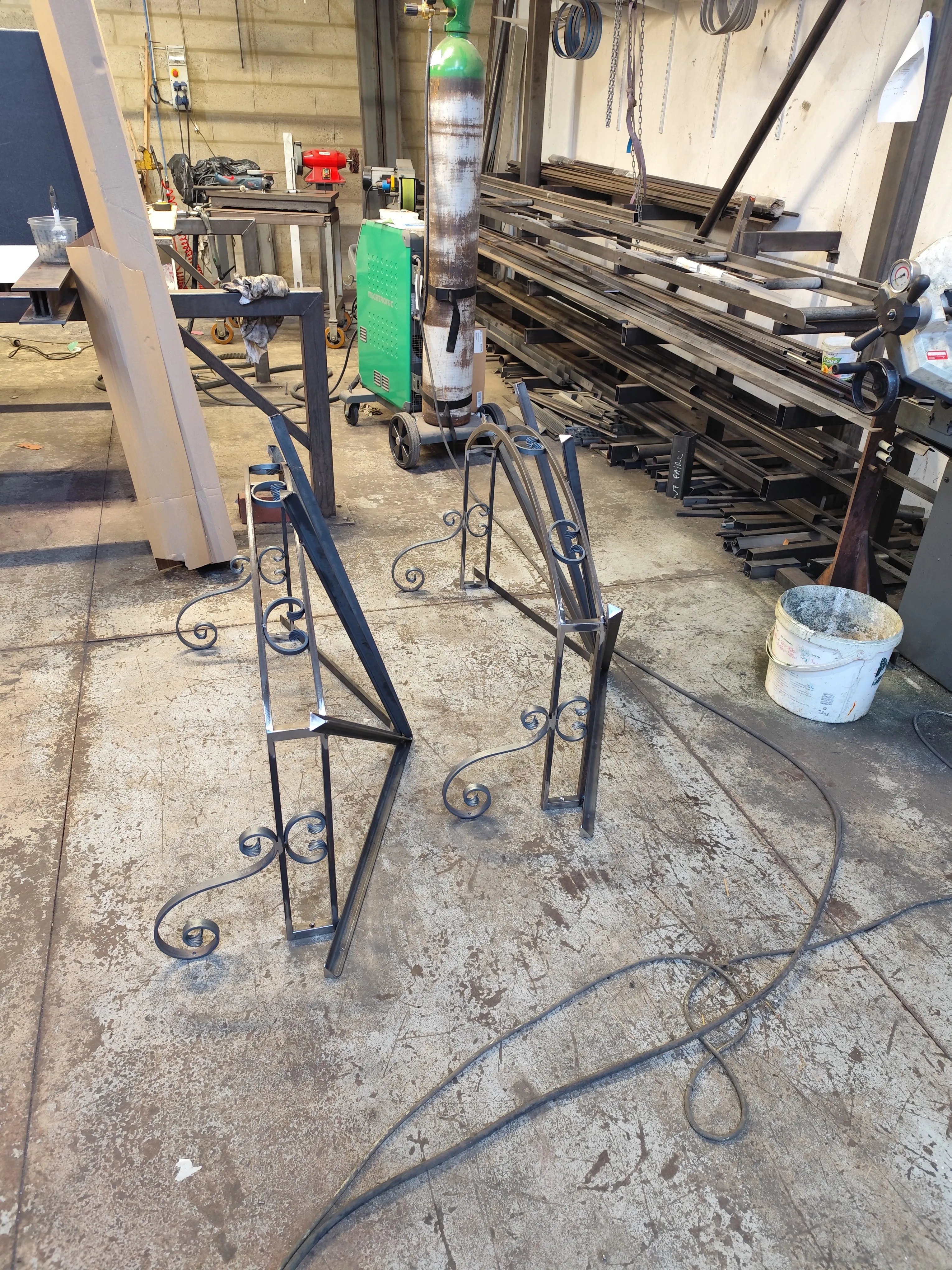 Deux structures latérales de marquises en acier avec motifs à volutes, posées au sol dans un atelier, illustrant la fabrication de pièces jumelles.