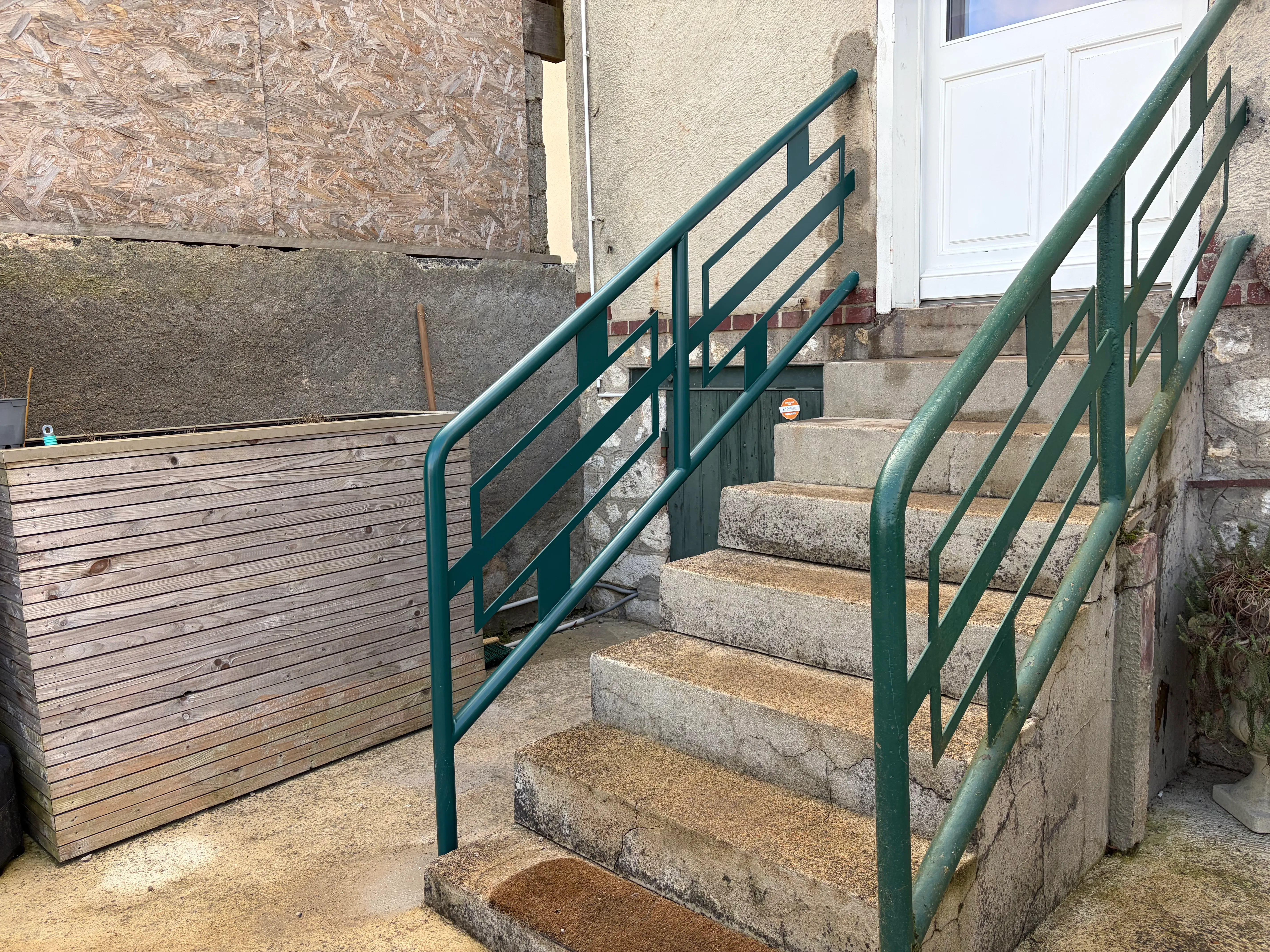 Rampe d'escalier extérieur en acier avec finition peinture polyuréthane verte anti-rouille. Garde-corps aux motifs géométriques rectangulaires fixé sur un escalier en béton menant à une entrée.