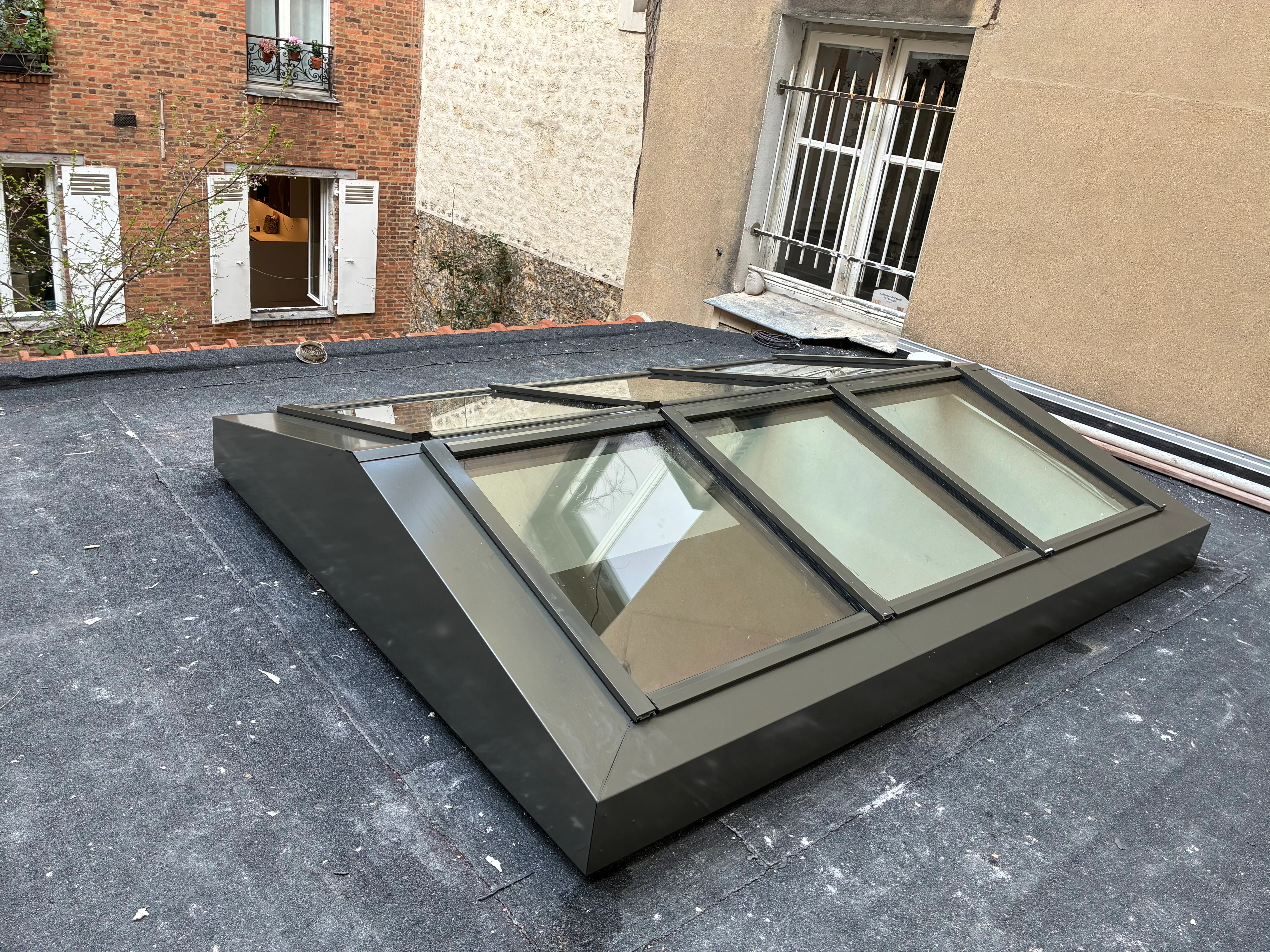 Vue d'ensemble d'une nouvelle verrière de toit à double pente en acier sombre, installée sur un toit terrasse parisien entouré de façades en briques et pierres.