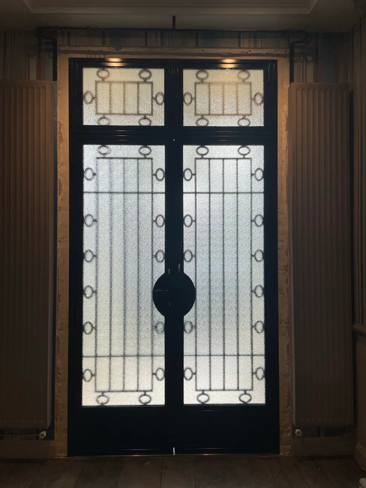 Vue intérieure d'une porte ou cloison métallique vitrée, révélant ses vitrages texturés lumineux, son armature noire structurée et sa grande serrure centrale